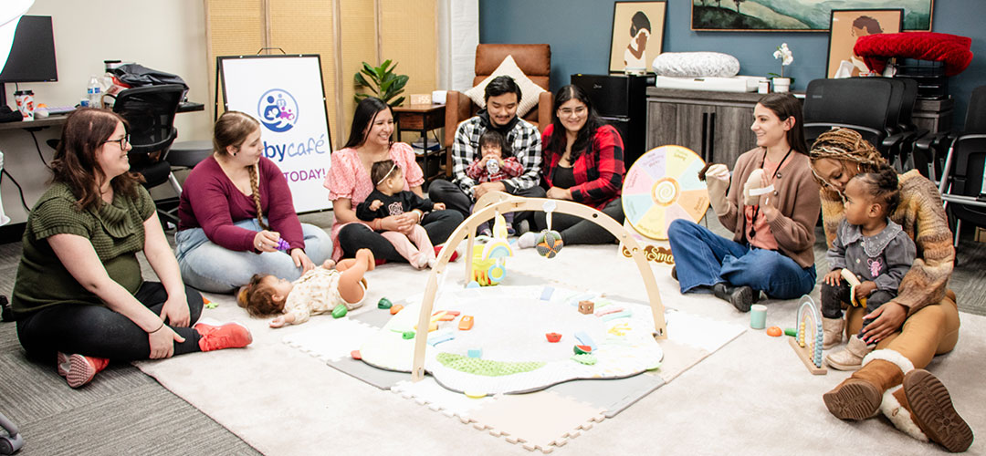 A group of adults and children sit in a circle at Baby Café surrounded by toys and activities.