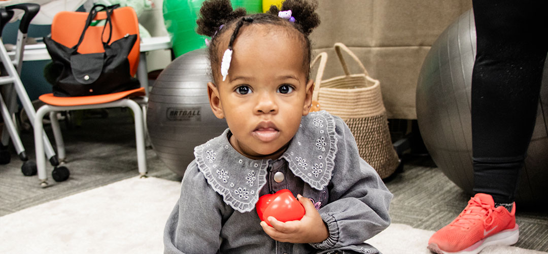 Small girl child with pigtails holds a red apple in a playroom.