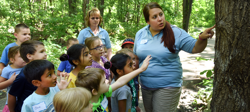 A guide points to a spot on a tree trunk while teaching a group of elementary school students in the woods.
