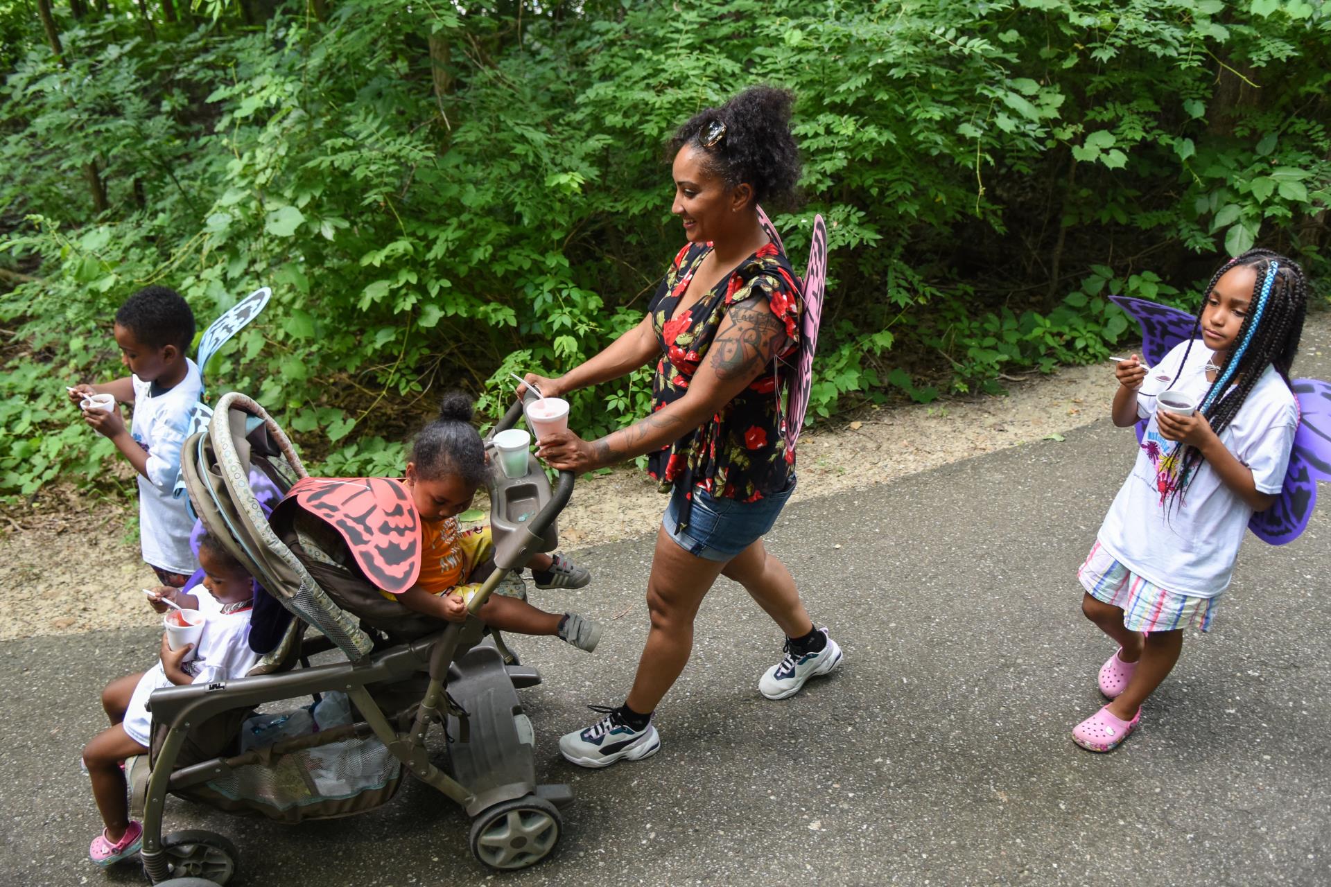 A smiling woman pushes two children in a stroller while a third child walks alongside.
