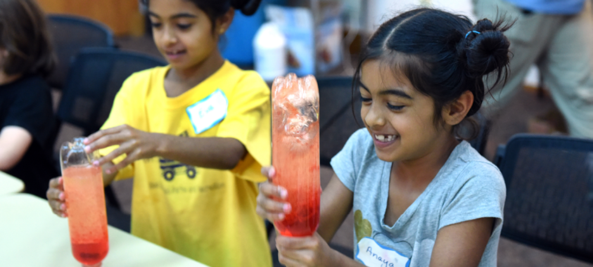Two smiling children conduct an experiment with a large bottle containing orange and red fluid.