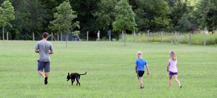 Two children and an adult walk in a field with their black dog at the Lyon Oaks dog park
